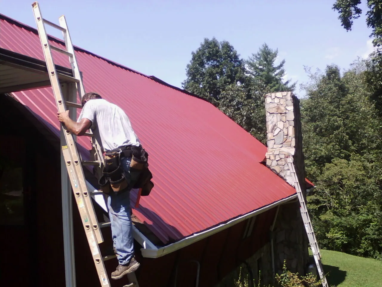 Roofer on a ladder beside a red metal roof and stone chimney — Soffit & Fascia Repair by Roofing Brothers in Washington